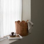 Handgjord korg i naturfärgad rotting med vågig kant placerad på fönsterbräda tillsammans med borstar och textilier.