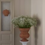 Naturfärgad rottingurna med vita blommor på piedestal i ljus inomhusmiljö.