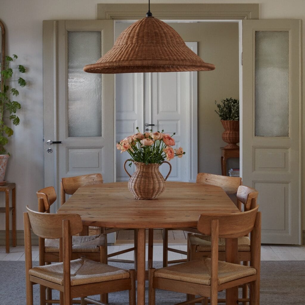Vas i rotting med blommor på matbord i varmt inrett rum med trästolar och rottinglampa.