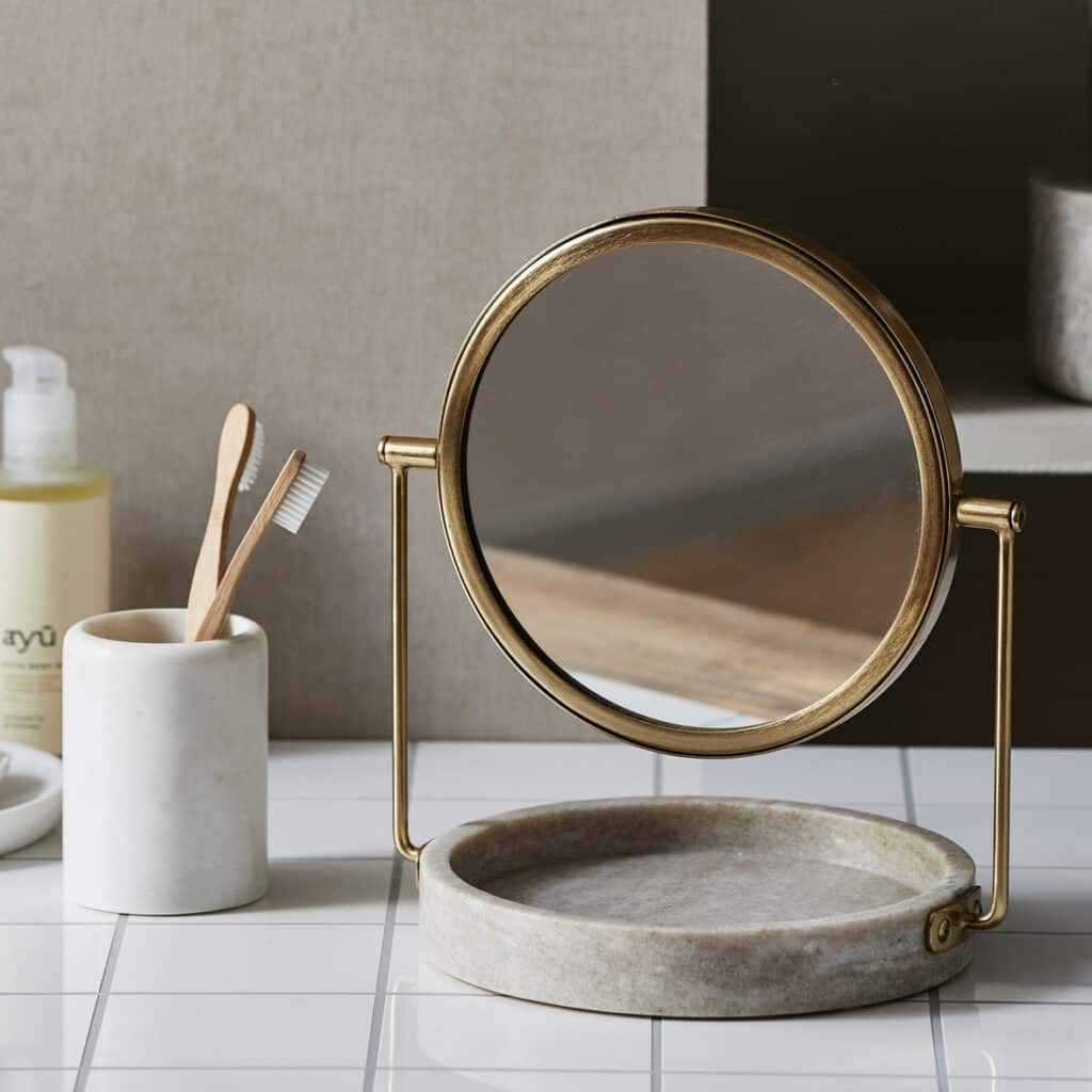 Rund bordsspegel i antik mässing på stenskiva, placerad på vit kakelyta i badrum tillsammans med tandborstmugg i keramik och trädetaljer.
