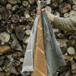 FIGARO handdukar i beige, rökt ek och grönt hålls framför vedstapel