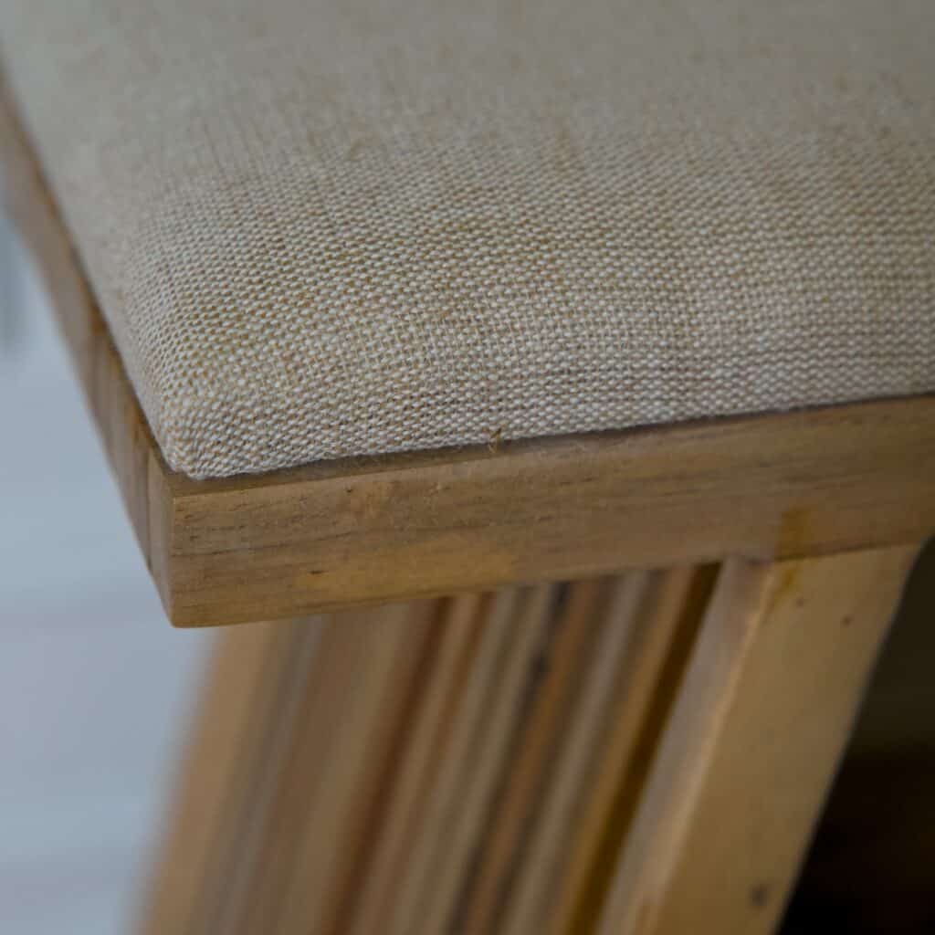Närbild på pall i teak med beige tygsits som visar vävd textur, naturlig träkant och ribbad träkonstruktion.