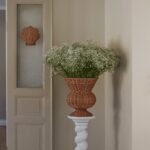 Rottingurna med vita blommor på piedestal och snäckformad rottingdekoration på dörr.