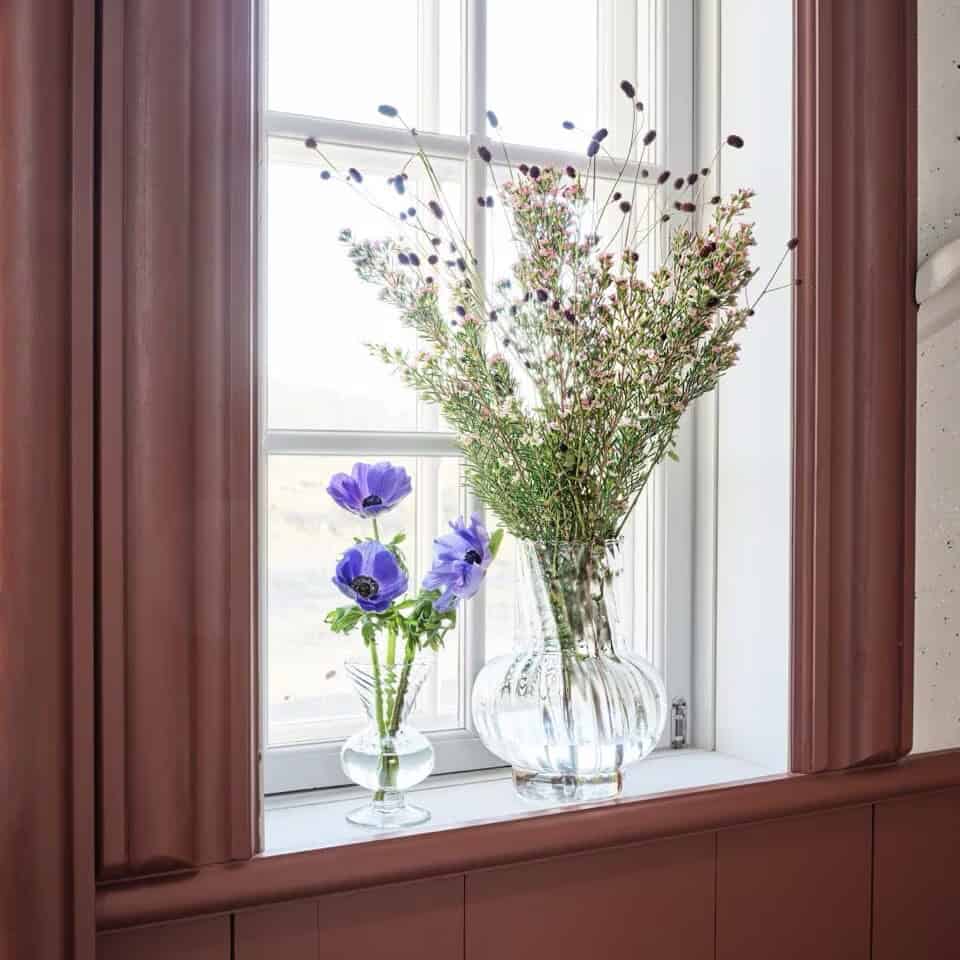 Glasvas Annie i klart glas med lila blommor i ett fönster.