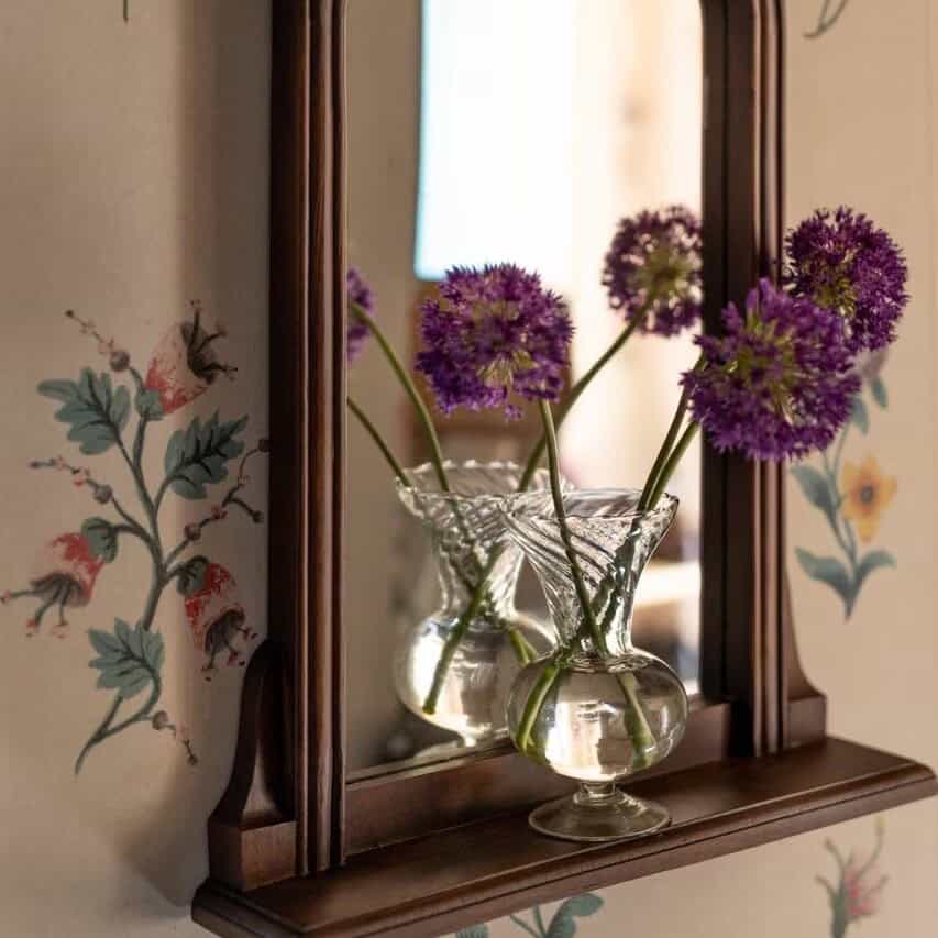 Glasvas Annie i klart glas med lila blommor framför en brun spegel.
