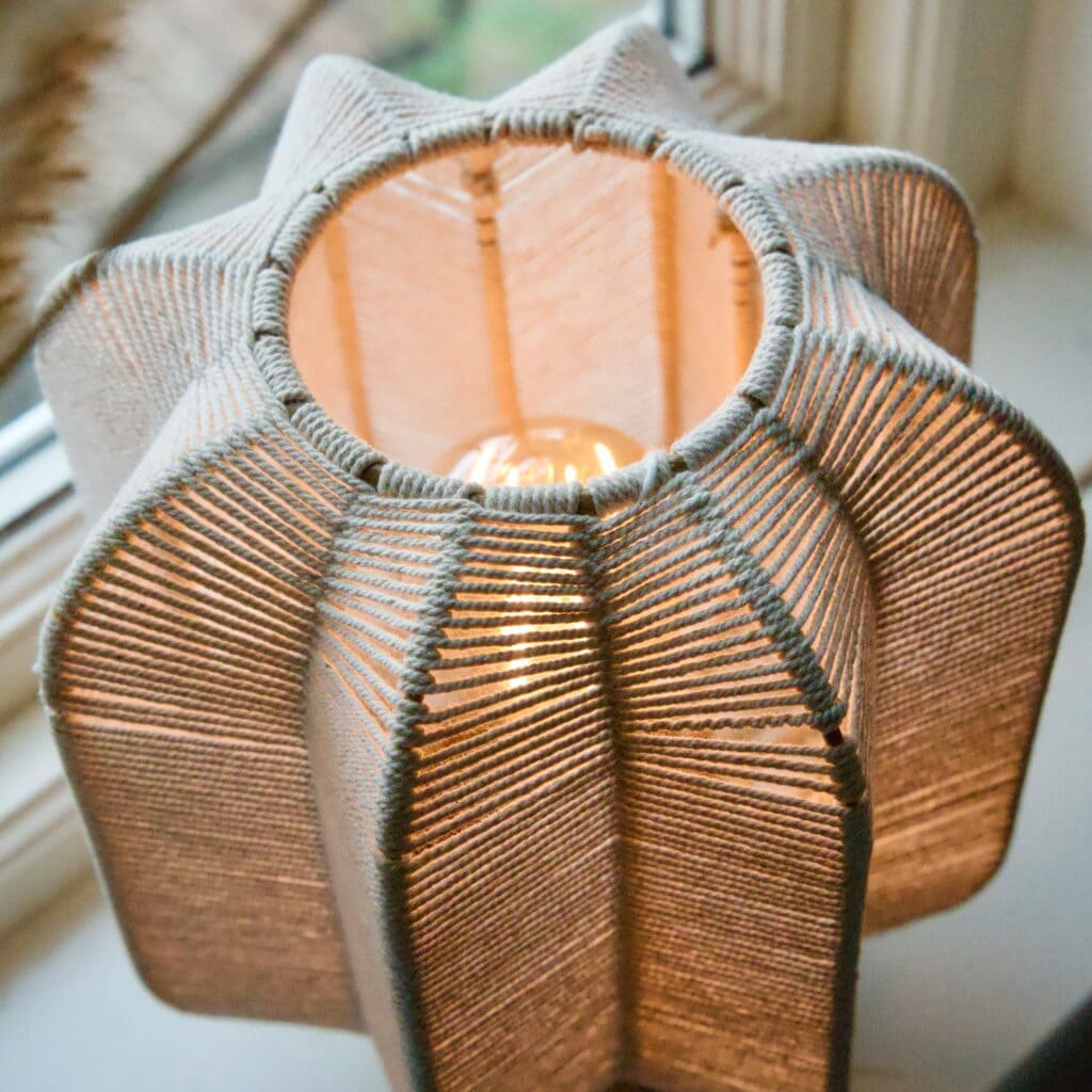 Närbild ovanifrån på tänd bordslampa med flätad skärm i offwhite bomullsrep och varmt ljus.
