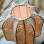 Närbild ovanifrån på tänd bordslampa med flätad skärm i offwhite bomullsrep och varmt ljus.