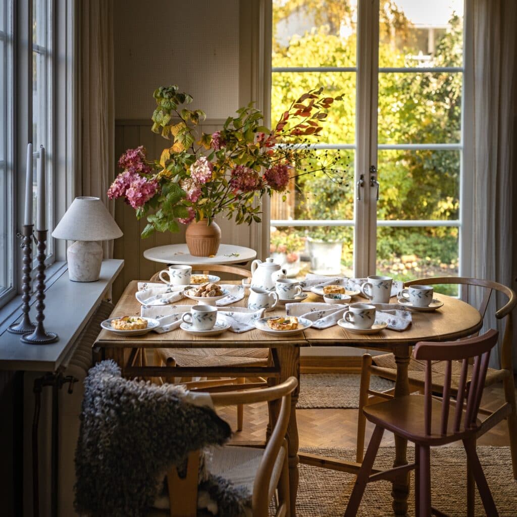 Höstdukning med kaffekoppar, små äppelpajer och blommor på ett träbord vid ett soligt fönster.