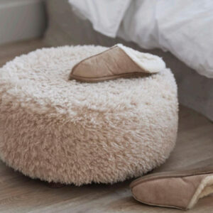 Rund sittpuff Leane i beige fluffig teddy placerad bredvid en säng med vita sängkläder och ett par tofflor.
